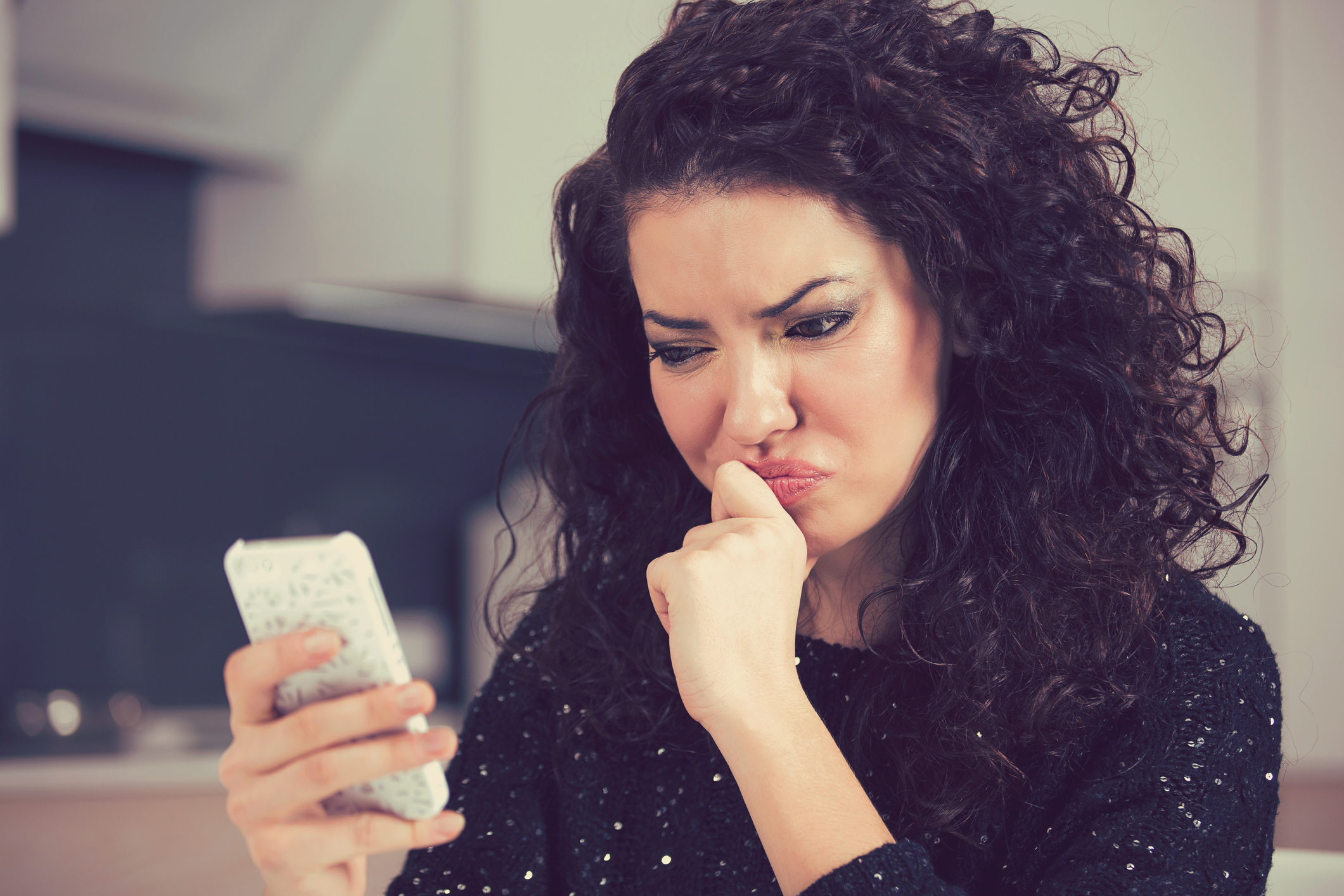 A woman with curly dark hair and light skin is looking at her smartphone with a displeased and frustrated expression. She is wearing a black, sequined sweater and is biting her thumb in contemplation. The background features a modern kitchen with neutral-colored cabinets.