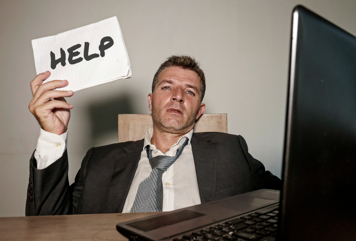 Man frustrated behind computer and desk holding help sign.